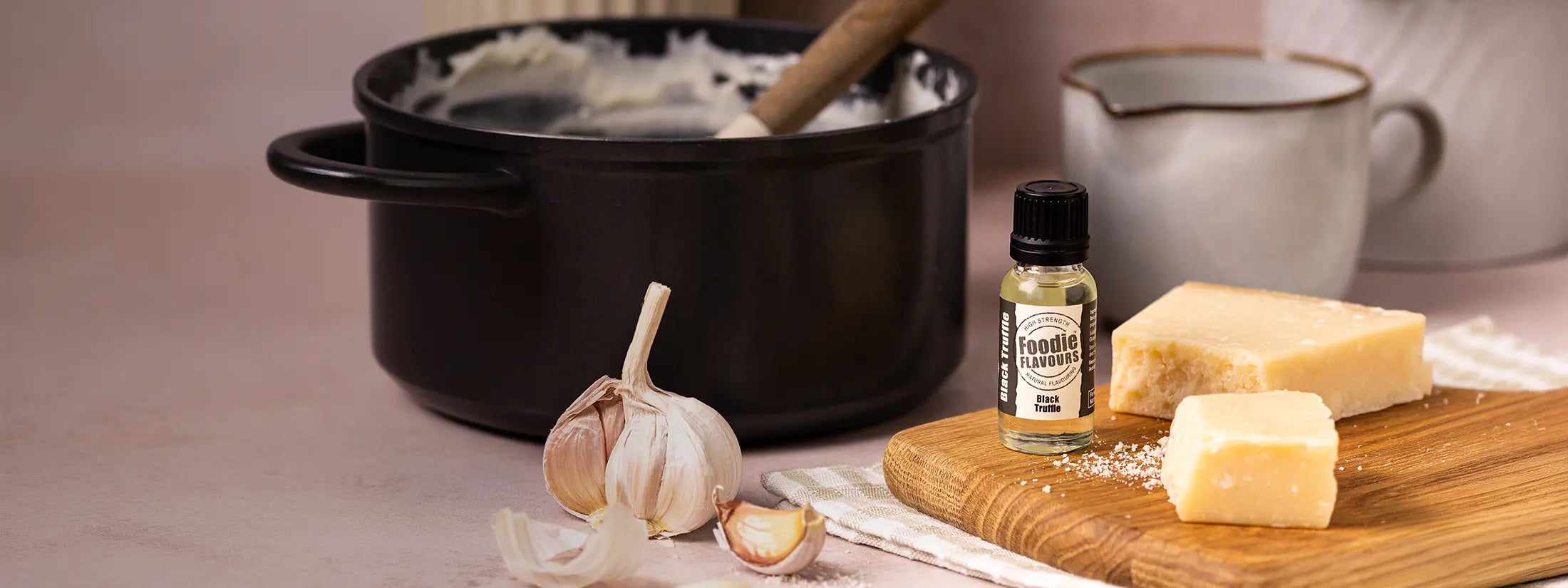 Black pot with ingredients on a wooden board, including garlic, cheese, and a bottle of Black Truffle Natural Flavouring.