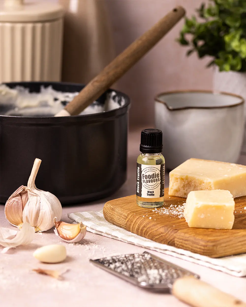 Small bottle of black truffle food flavouring on a wooden cutting board with garlic, cheese, and a pot in the background.