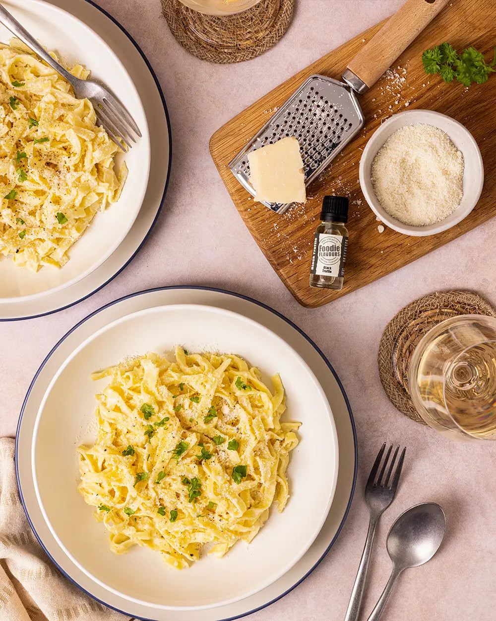 Two plates of pasta with a grater, cheese, and a bottle of black truffle flavouring on a table.