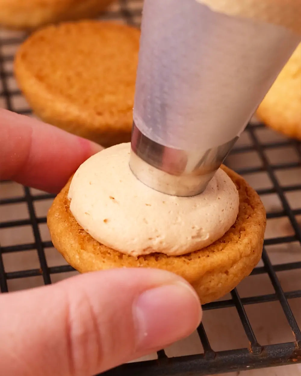 Person filling a cookie with cream using a piping bag on a cooling rack.