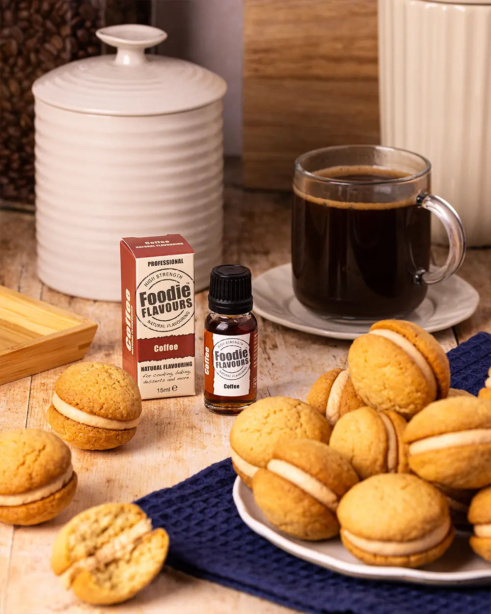 Coffee-themed setting with coffee kisses biscuits, coffee, and Foodie Flavours coffee flavouring on a wooden surface.