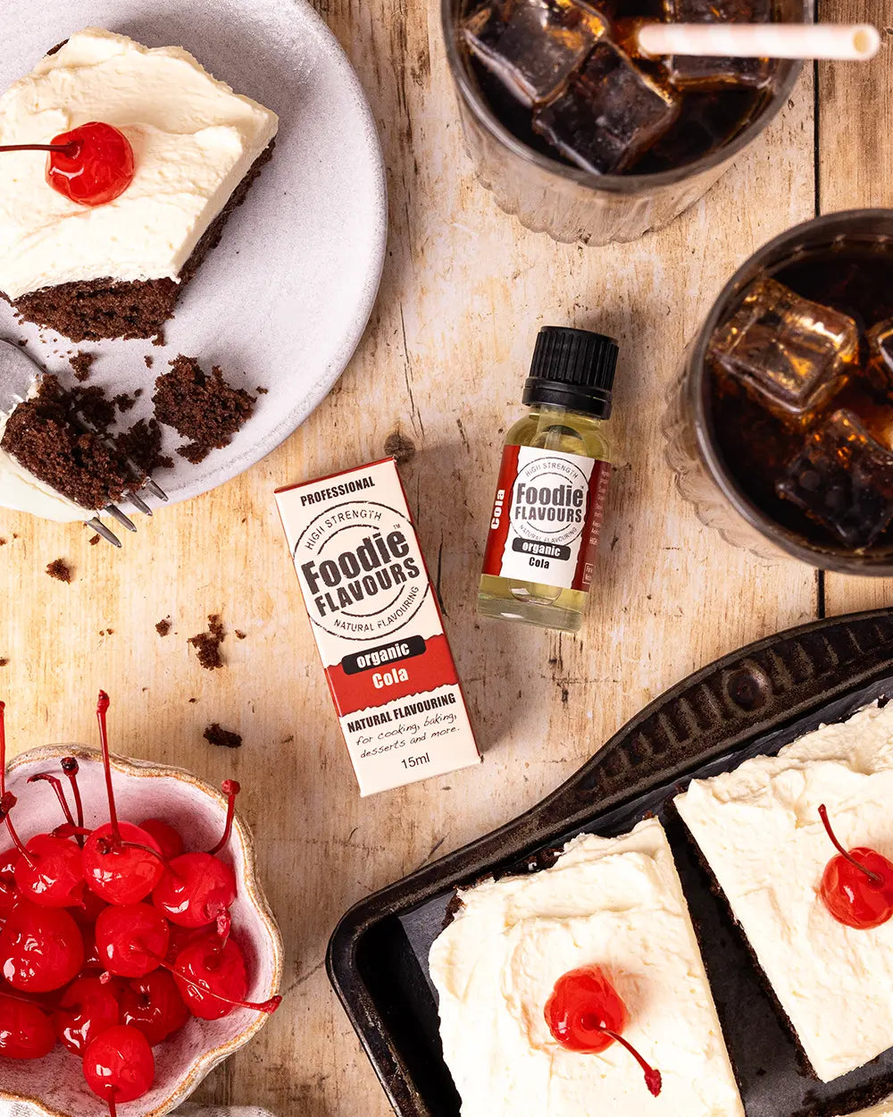 Chocolate cake with whipped cream and cherries, bottle of Foodie Flavours organic cola, and bowls of cherries on a wooden surface.