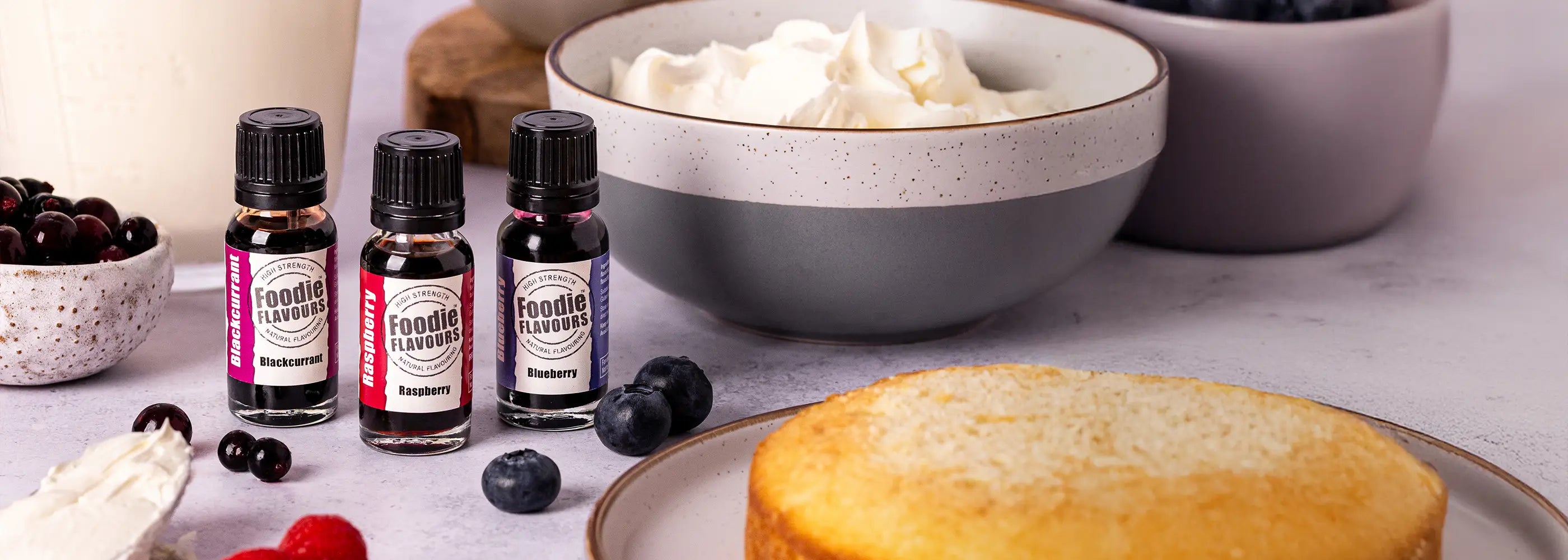 Bowl of whipped cream, cake, and bottles of foodie flavours blackcurrant, raspberry and blueberry natural food flavourings on a table.