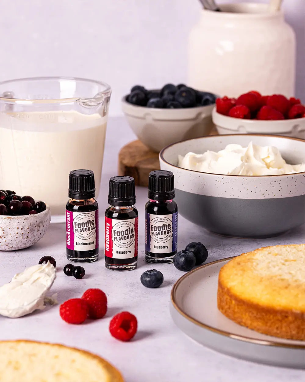 Three small bottles of foodie flavours: Blackcurrant, Raspberry and Blueberry. With berries, cream, and cake on a light background