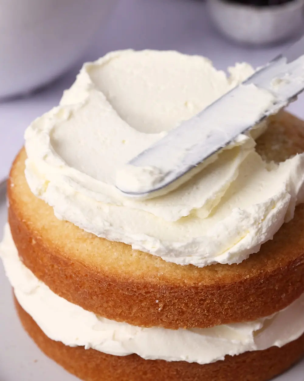Layered cake being frosted with white cream using a spatula.
