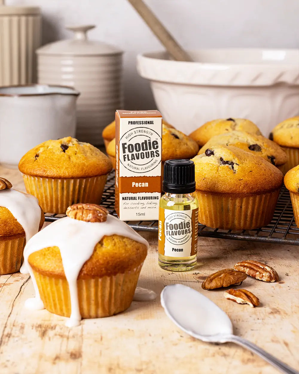 Pecan Muffins with icing, a bottle of Foodie Flavours pecan essence, and pecans on a wooden surface.