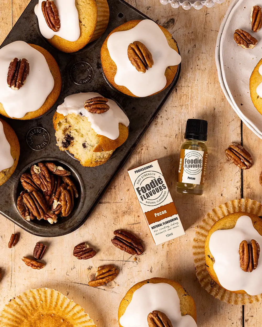Muffins with white icing and pecans on a wooden surface with a bottle of foodie flavours pecan flavouring.