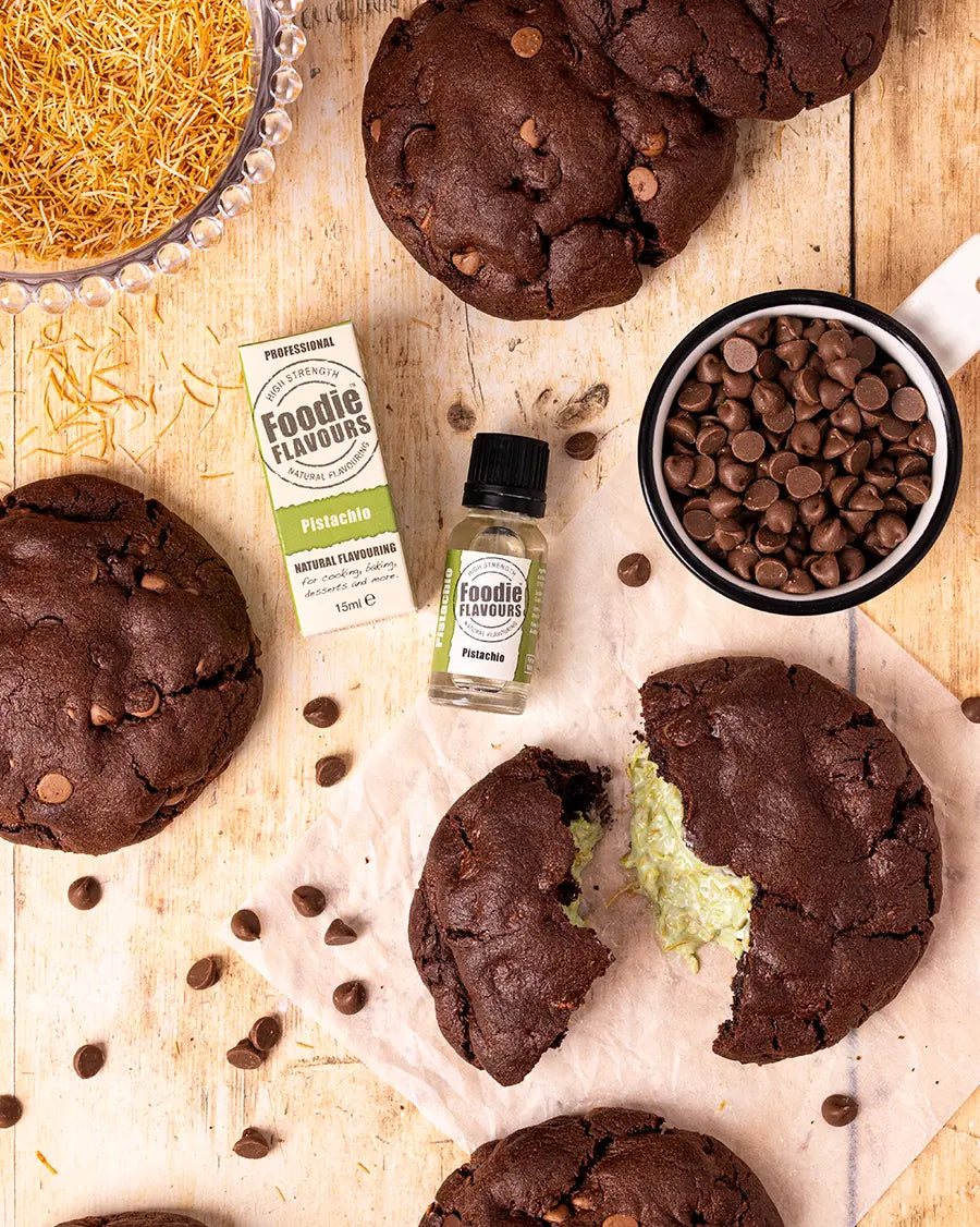 Chocolate cookies with a green Dubai chocolate filling, Foodie Flavours pistachio flavouring bottle, and chocolate chips on a wooden surface.