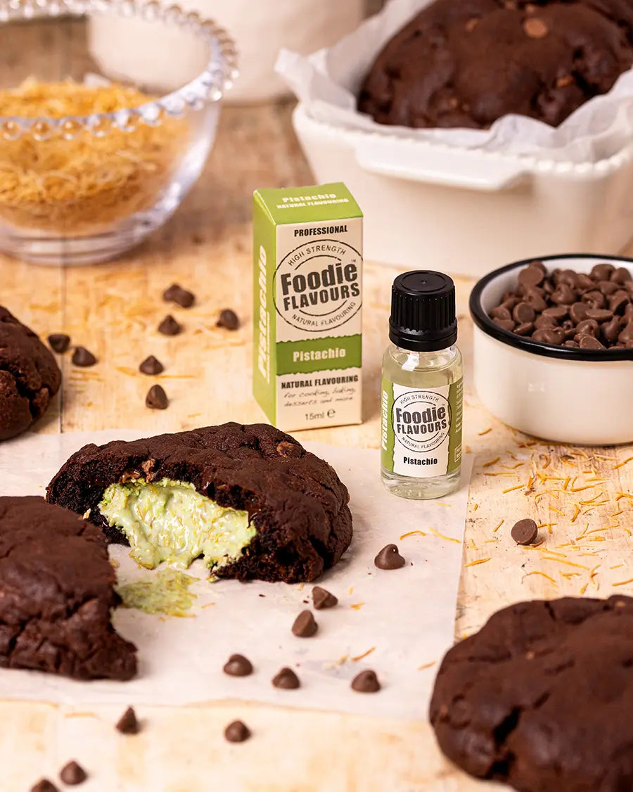 Chocolate cookies with a green Dubai chocolate filling, Foodie Flavours pistachio flavouring bottle, and chocolate chips on a wooden surface.