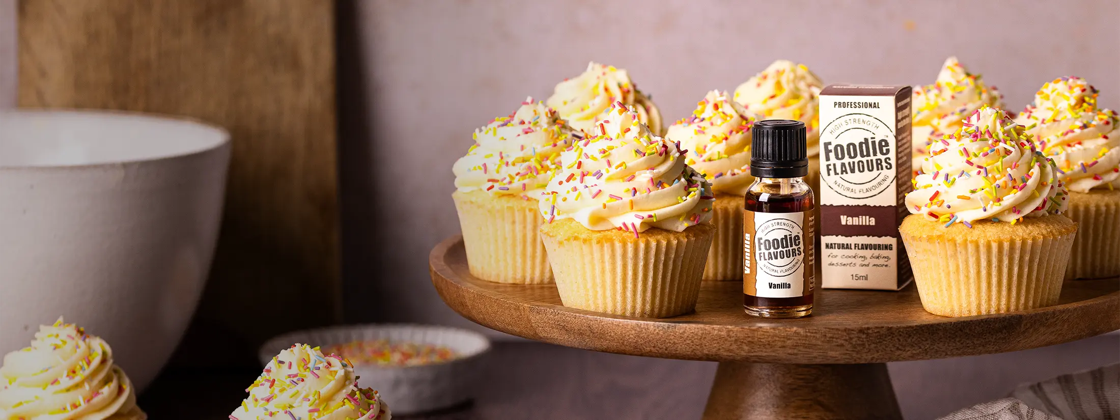 Glass bottle of Foodie Flaovurs' Vanilla Natural Food Flavouring on a wooden cake stand with vanilla flavoured cupcakes topped with buttercream and sprinkles.