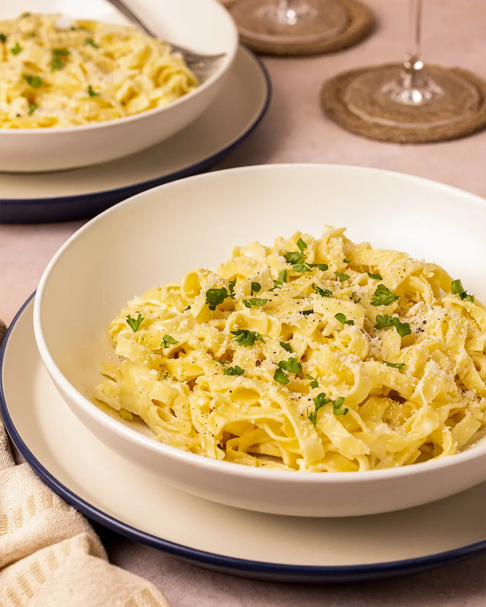 Two bowls of pasta with a light sauce and green herbs on a beige tablecloth.