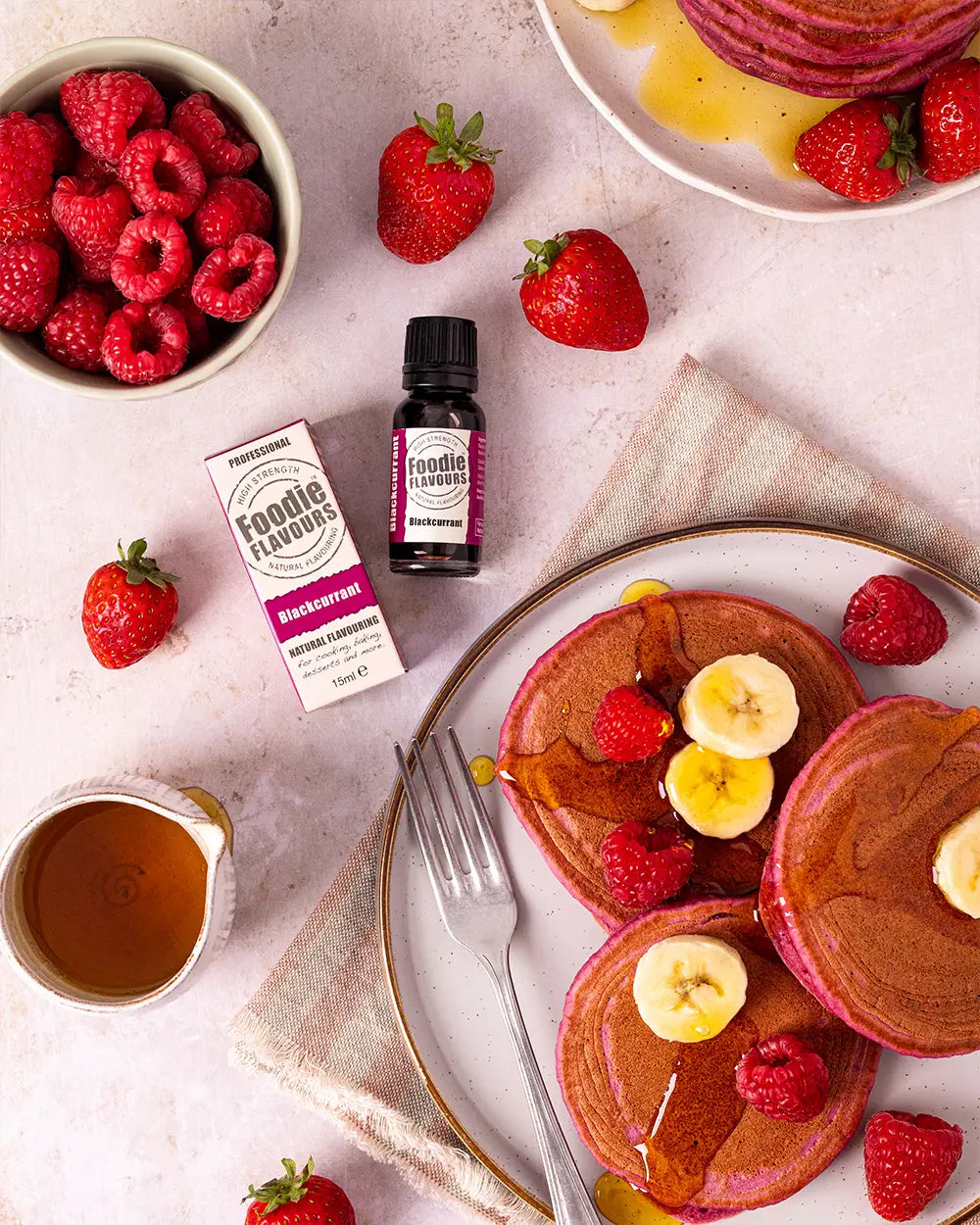 Pancakes with fruit on a plate, surrounded by raspberries, strawberries, and a bottle of foodie flavours blackcurrant essence.