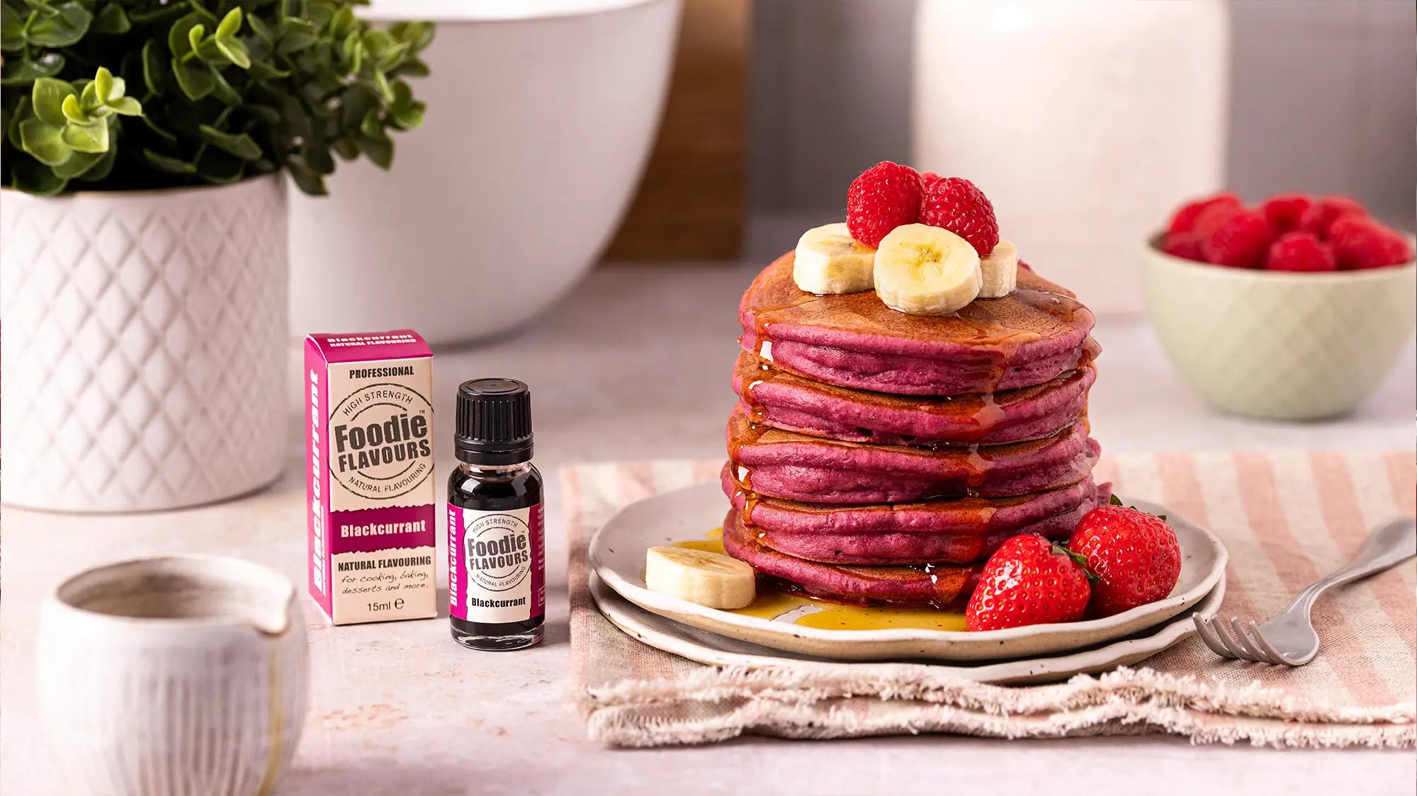 Stack of pink pancakes with berries and bananas on a plate, next to a box and bottle of blackcurrant food flavouring.