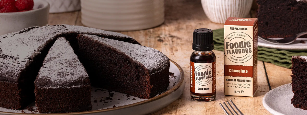 A bottle and box of Foodie Flavours Chocolate flavouring placed beside a sliced chocolate cake on a wooden table, with additional cake slices and bowls in the background.
