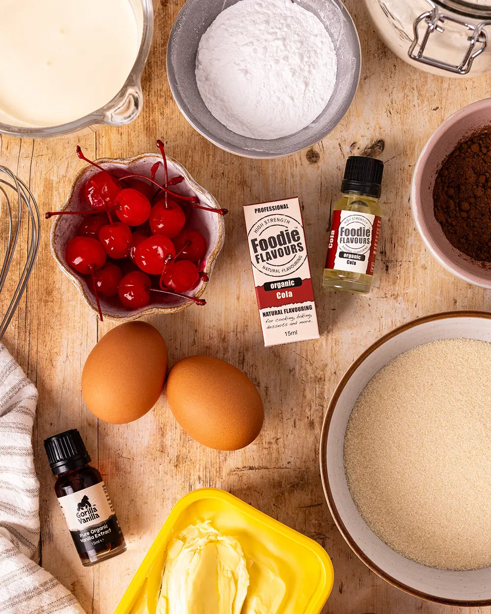 Ingredients and cola flavouring and vanilla extract for baking on a wooden surface, including eggs, flour, cherries.