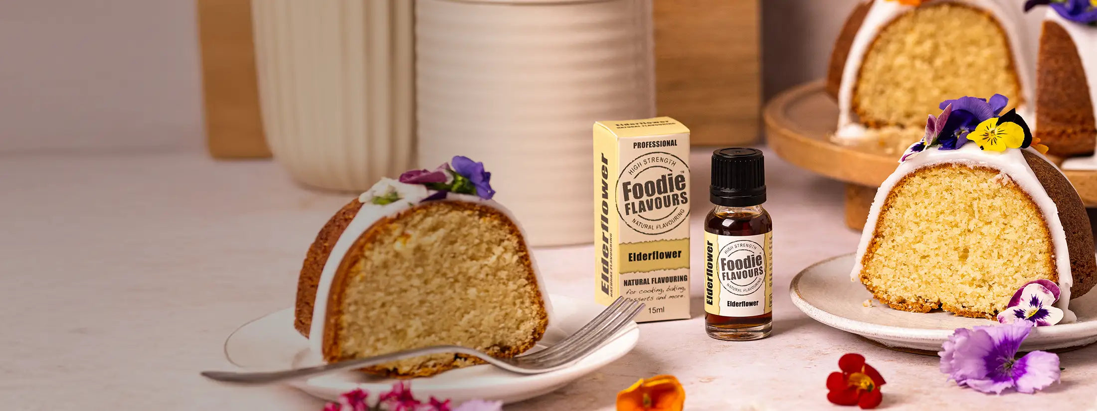 A small bottle and box of Foodie Flavours Elderflower Extract is displayed on a tabletop beside slices of glazed cake, with flowers scattered around and a softly blurred kitchen-like background.