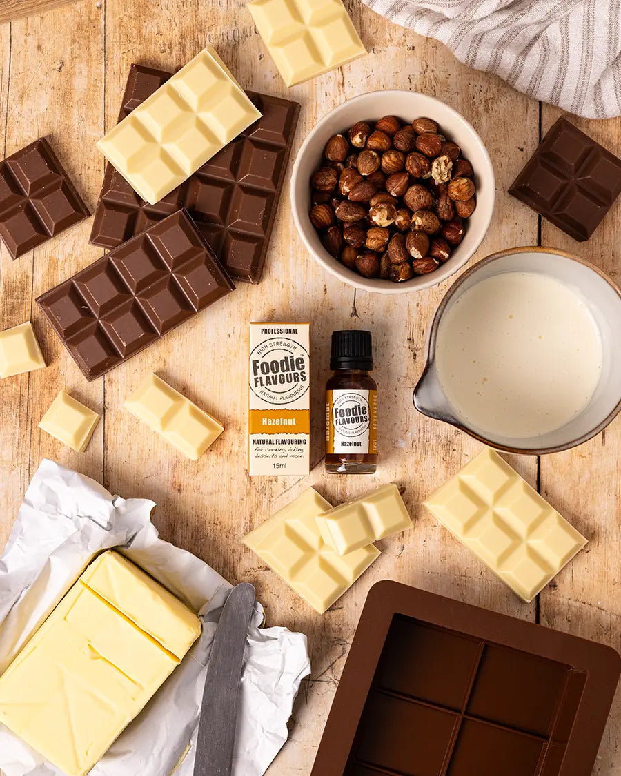 Assorted chocolate bars, a bowl of hazelnuts, a bottle of foodie flavours hazelnut food flavouring, and a container of cream on a wooden surface.