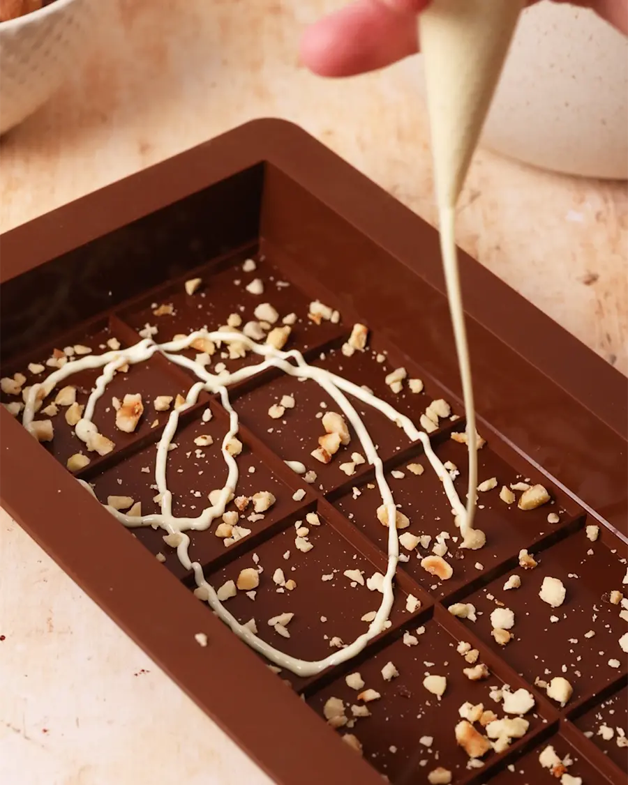 Chocolate bar mould being prepared with white drizzle and nuts being decorated with a piping bag.