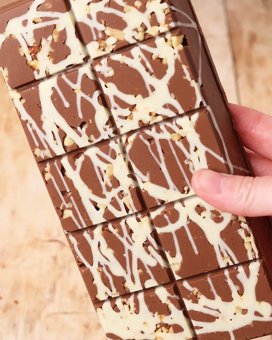 Chocolate bar with white drizzle and nuts held by a hand on a wooden surface