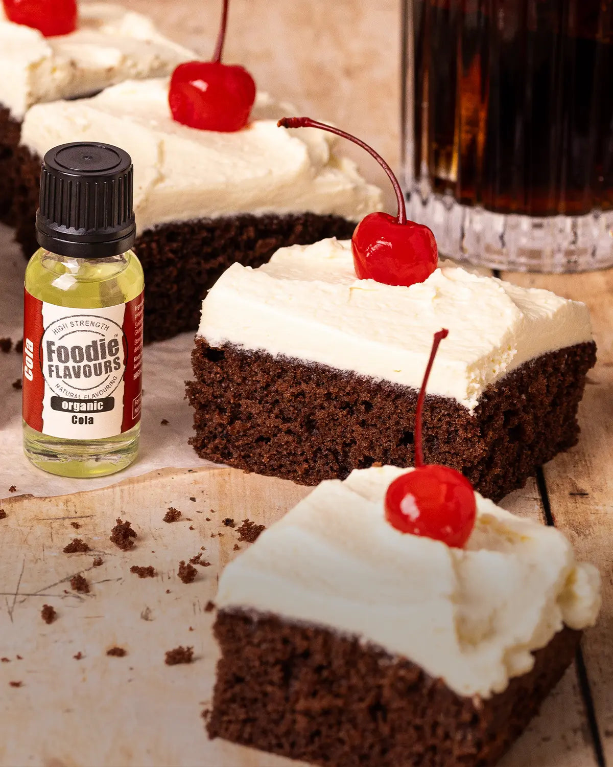 Chocolate traybake cake with white vanilla cream topping and a cherry on top, next to a bottle of foodie flavours cola food flavouring.