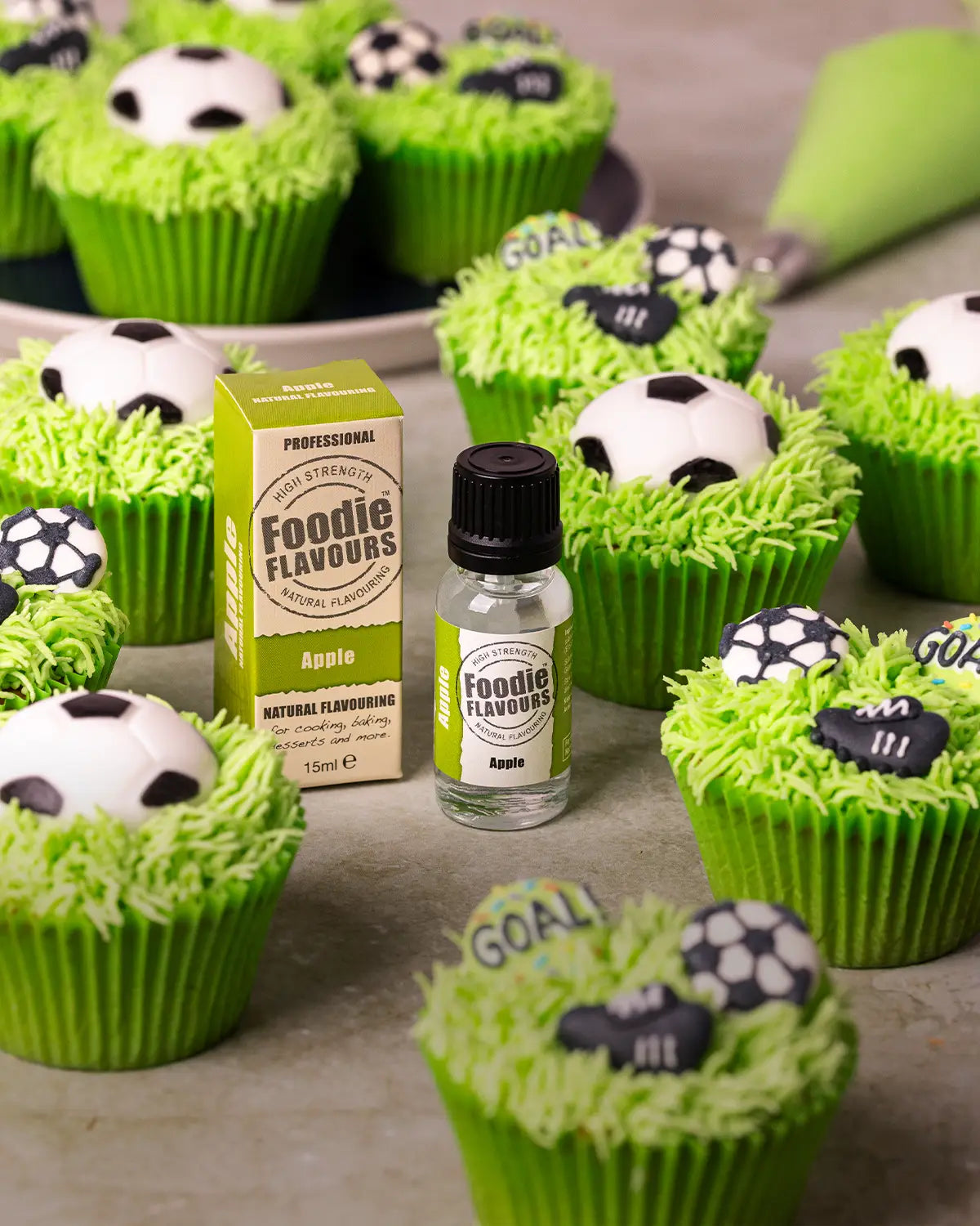 A bottle and box of Foodie Flavours Apple natural flavouring surrounded by decorated cupcakes with bright green frosting, football toppers, and football boots