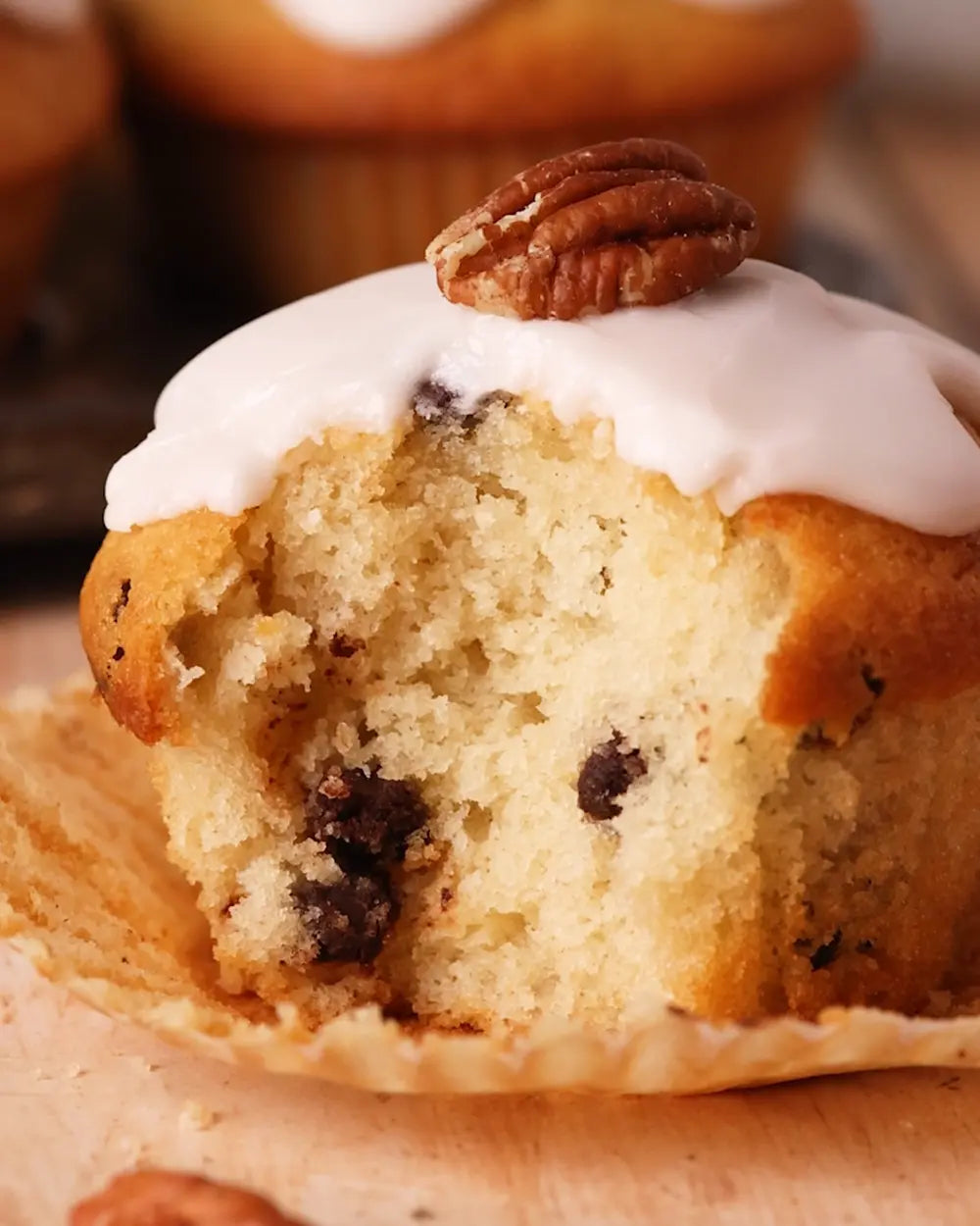 Half-eaten pecan muffin with white frosting and a pecan on top, with other muffins in the background.