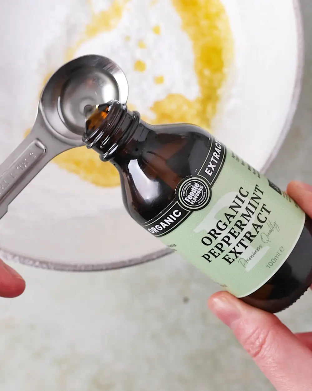 Bottle of Organic Peppermint Extract being poured into a mixing bowl with a measuring spoon.