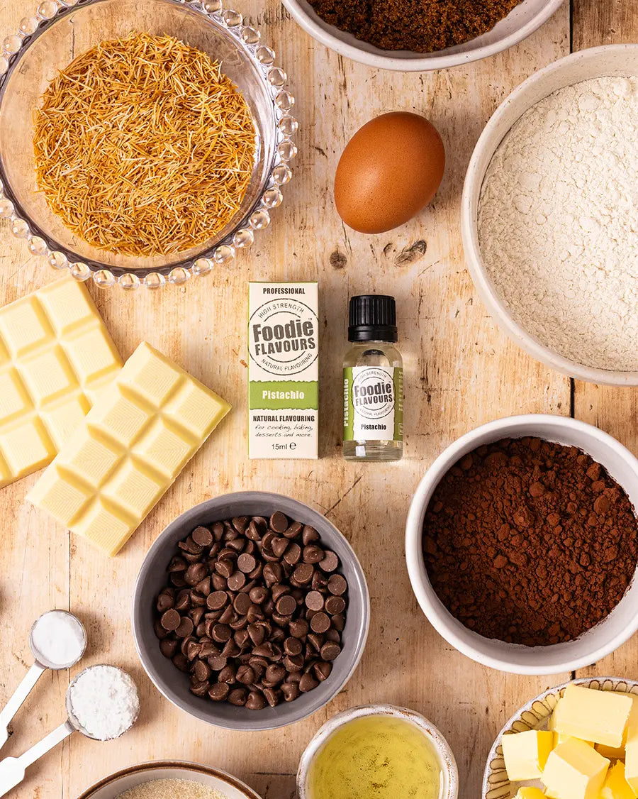 Assorted baking ingredients including chocolate chips, flour, pistachio flavouring and butter on a wooden surface.