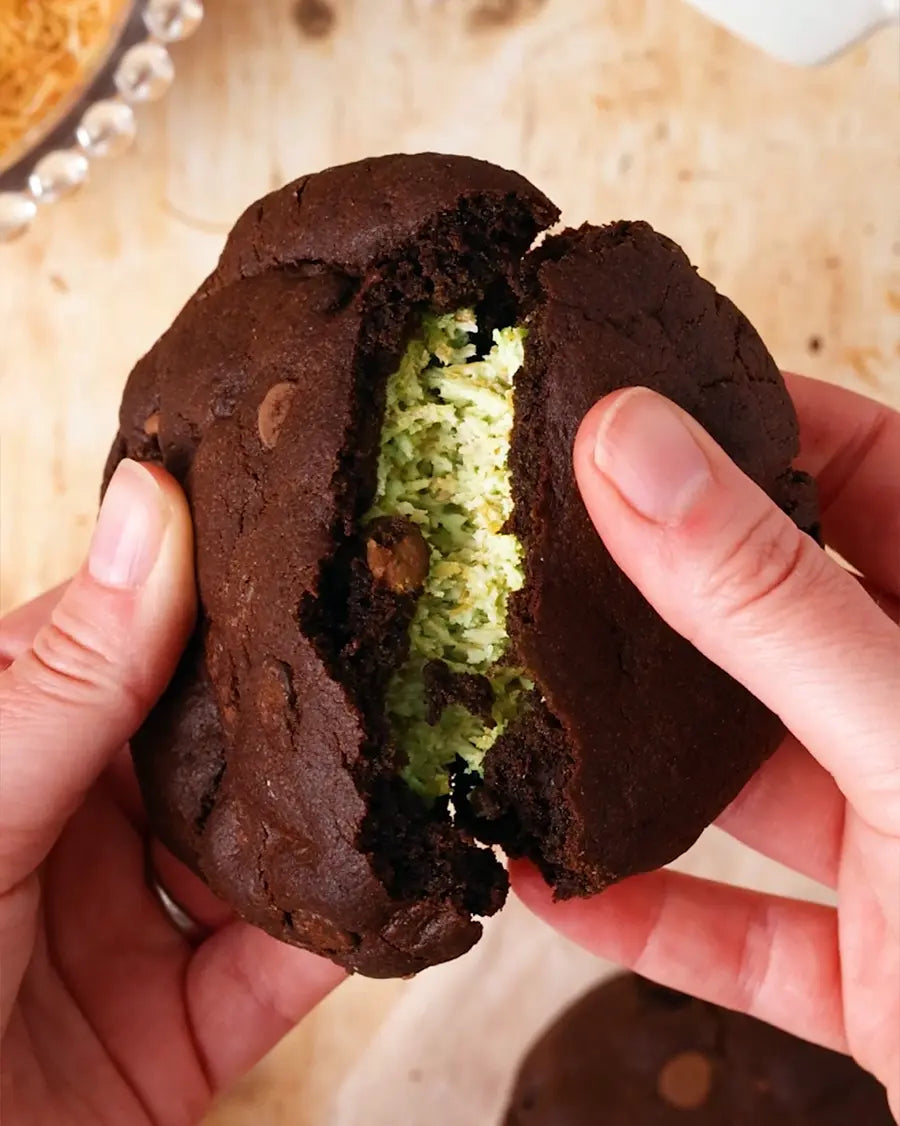 Chocolate cookie with green Dubai Chocolate filling held between two hands on a wooden surface.