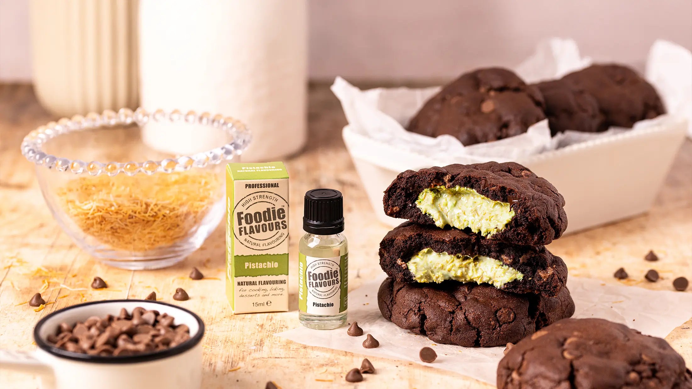 Chocolate cookies with a green Dubai chocolate filling, Foodie Flavours pistachio flavouring bottle, and chocolate chips on a wooden surface.