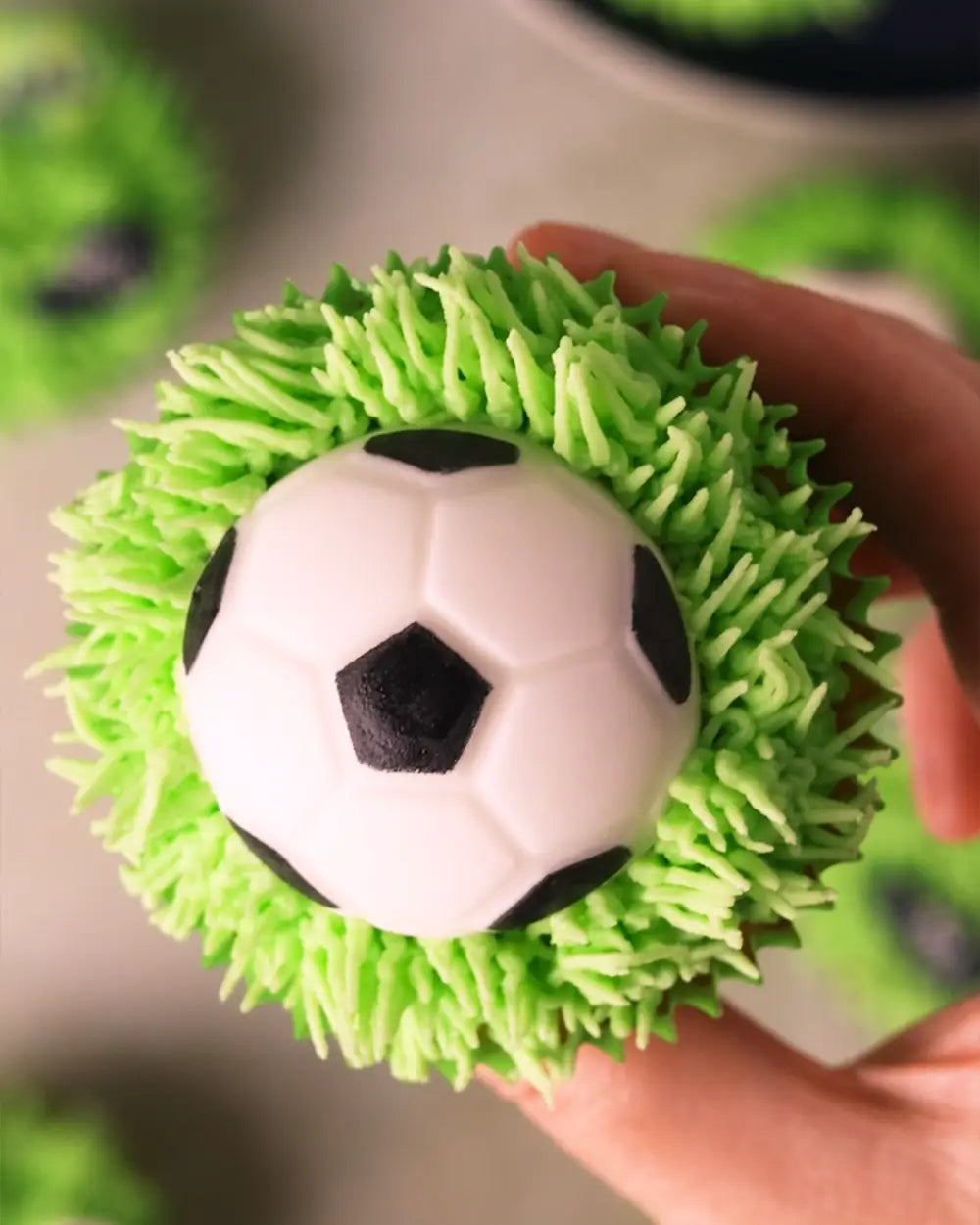 A close-up of a cupcake topped with bright green whipped frosting and a small white football decoration, held by a hand against a blurred background.