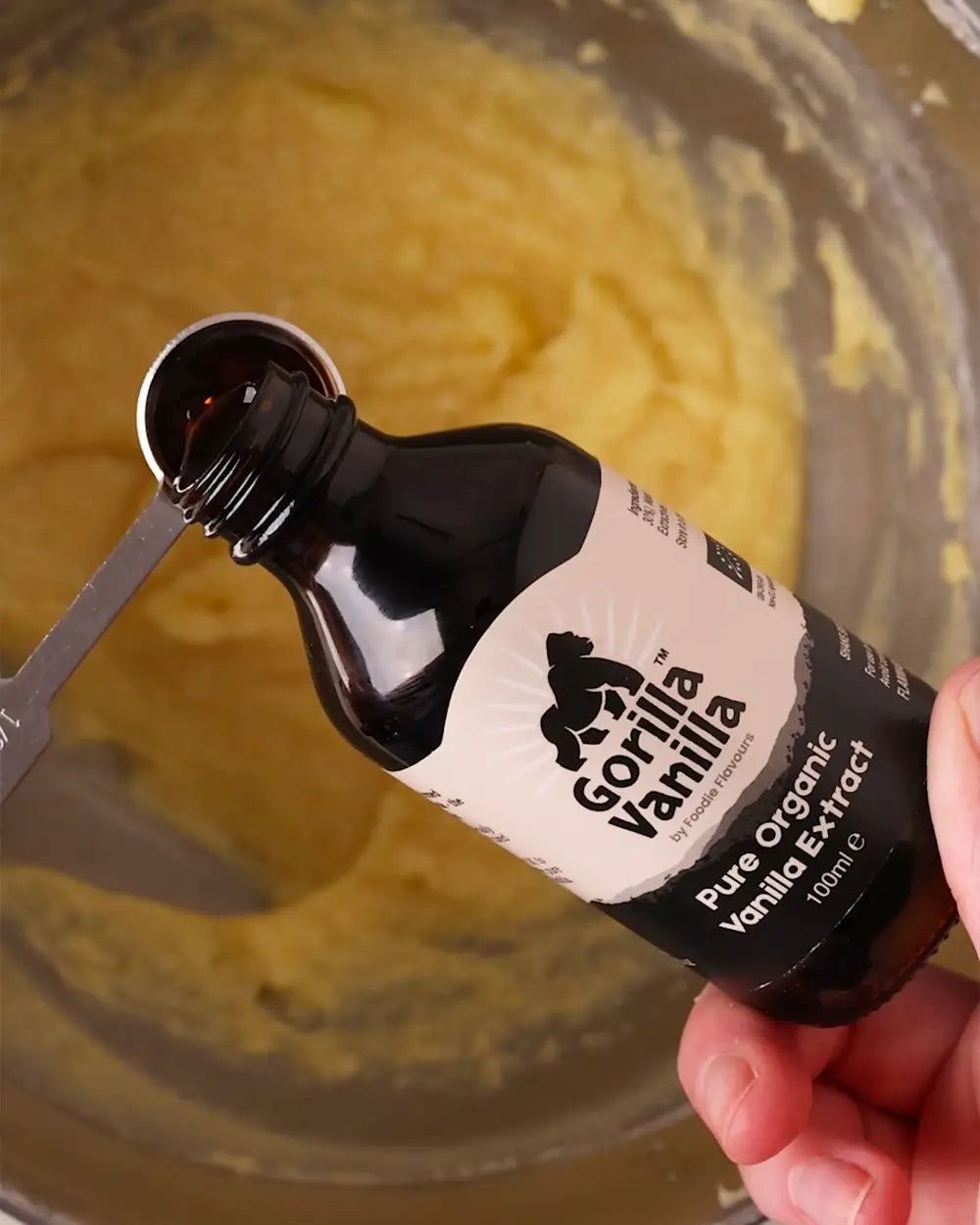 A hand holding a bottle of Gorilla Vanilla Pure Organic Vanilla Extract over a bowl of yellow batter, with a metal measuring spoon visible.