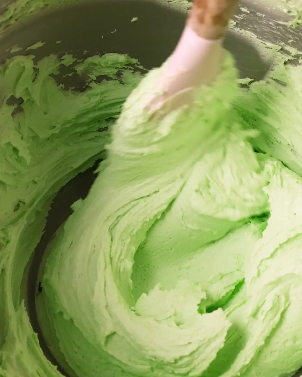Light green apple flavoured whipped frosting or cream being stirred with a spatula inside a dark mixing bowl.