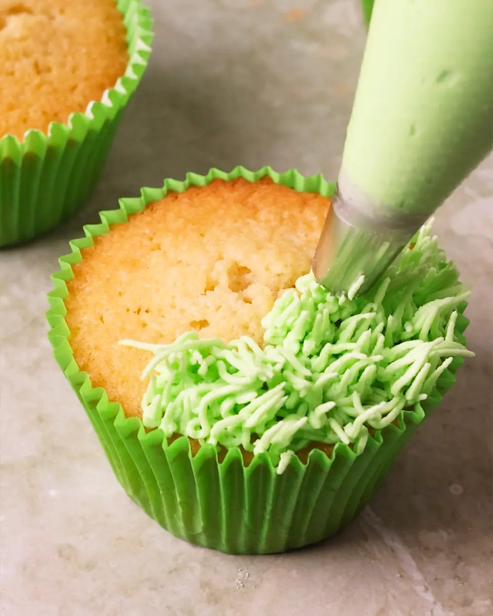 A close-up of a green cupcake liner containing a baked cupcake, with bright green frosting being piped onto the top and side. Another cupcake is partially visible in the background on a neutral surface.