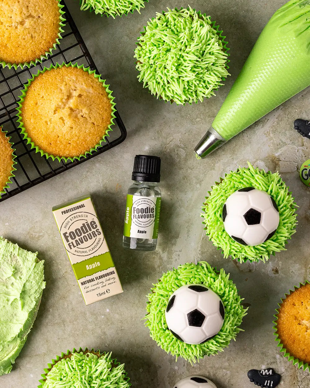 A styled flat-lay of green-frosted cupcakes, some topped with football decorations, alongside a piping bag, a bottle of Foodie Flavours apple on a grey surface.